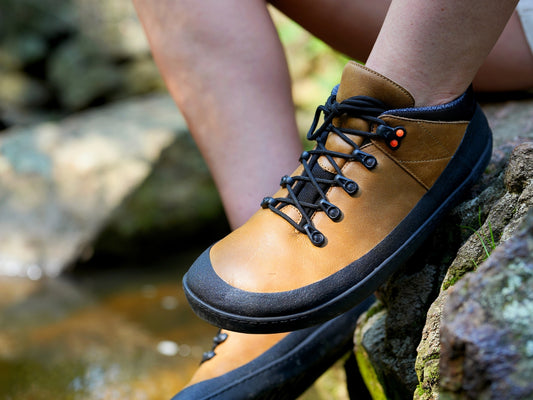 Brown barefoot hiking shoes with black soles worn by a person sitting on a rock near water.